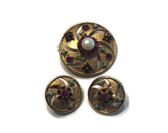 Peut inclure: Une broche dorée et une paire de boucles d'oreilles assorties présentant un motif circulaire avec des découpes en spirale. Chaque pièce est ornée d'une perle blanche centrale, de pierres rouge foncé et de petites perles turquoise.