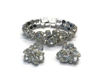 Op de afbeelding: Een zilverkleurige metalen armband en een paar bijpassende clip-oorbellen, elk voorzien van heldere, gefacetteerde marquise- en rondgeslepen strass-steentjes in een clusterpatroon. De armband is voorzien van een veilige sluiting.