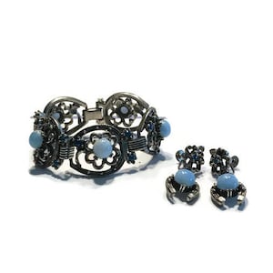 K&ouml;nnte beinhalten: Ein silberfarbenes Armband und ein passendes Ohrring-Set. Das Armband hat offene florale Designs mit hellblauen Cabochon-Steinen. Die Ohrringe haben ein &auml;hnliches Design mit kleineren blauen Steinen.