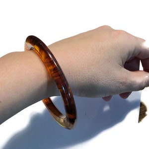 May include: A circular bangle bracelet featuring a translucent, polished finish with a marbled pattern of deep brown and amber tones. The rigid, smooth-surfaced accessory has a rounded profile and a glossy appearance that catches the light.