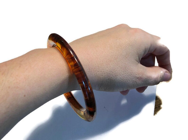 May include: A circular bangle bracelet featuring a translucent, polished finish with a marbled pattern of deep brown and amber tones. The rigid, smooth-surfaced accessory has a rounded profile and a glossy appearance that catches the light.