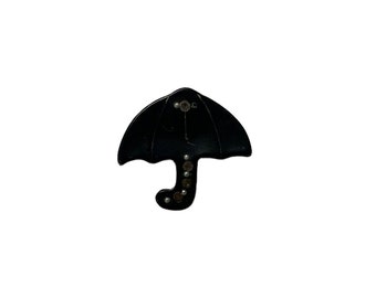 May include: A black umbrella-shaped brooch with a curved handle. The brooch has a glossy finish and is adorned with small, round, metallic accents. The umbrella's canopy is slightly curved, and the handle is a curved shape.