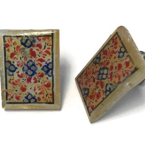 May include: A pair of square earrings featuring a floral pattern. The earrings have a cream-coloured base with a design of red, blue, and yellow flowers. The edges are outlined in gold. The earrings are set against a white backdrop.