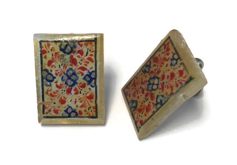 May include: A pair of square earrings featuring a floral pattern. The earrings have a cream-coloured base with a design of red, blue, and yellow flowers. The edges are outlined in gold. The earrings are set against a white backdrop.