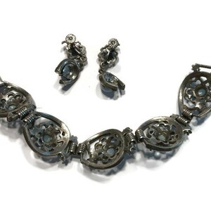 K&ouml;nnte beinhalten: Ein Vintage-Schmuckset aus silberfarbenem Armband und passenden Ohrringen. Das Armband hat verzierte, offene ovale Glieder mit floralem Design und kleinen, hellblauen Steinen. Die Ohrringe haben ein &auml;hnliches Design. Das Schmuckset wird auf wei&szlig;em Hintergrund pr&auml;sentiert.