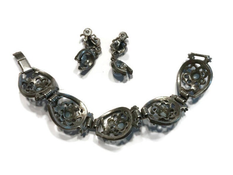 K&ouml;nnte beinhalten: Ein Vintage-Schmuckset aus silberfarbenem Armband und passenden Ohrringen. Das Armband hat verzierte, offene ovale Glieder mit floralem Design und kleinen, hellblauen Steinen. Die Ohrringe haben ein &auml;hnliches Design. Das Schmuckset wird auf wei&szlig;em Hintergrund pr&auml;sentiert.