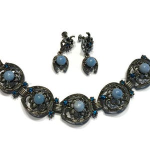 K&ouml;nnte beinhalten: Ein Vintage-Schmuckset mit Armband und passenden Ohrringen. Das Armband hat eine Reihe von verzierten, dunklen Metallgliedern, jedes mit einem zentralen hellblauen Stein und blauen Akzenten. Die Ohrringe haben ein Hufeisen-Design mit einem hellblauen Stein.