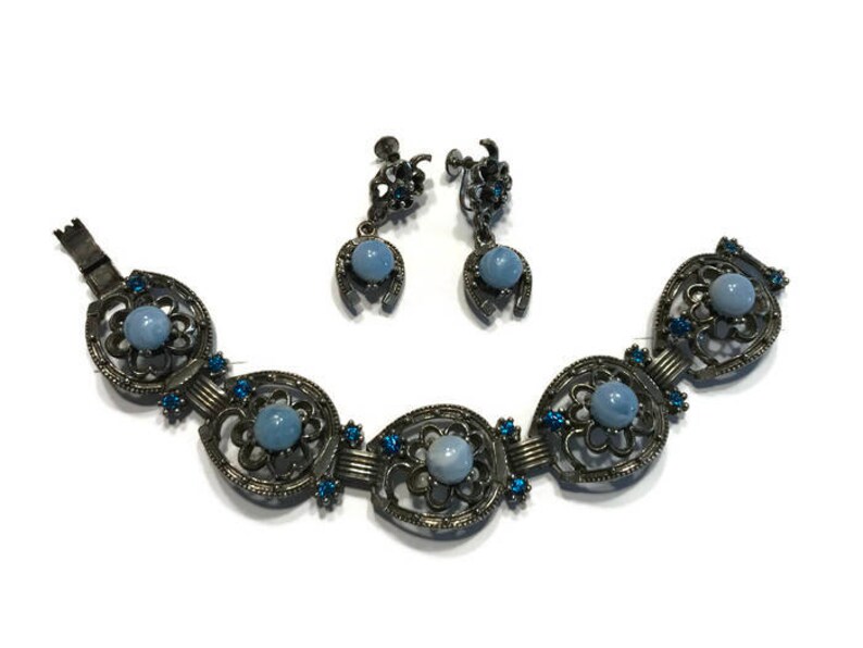K&ouml;nnte beinhalten: Ein Vintage-Schmuckset mit Armband und passenden Ohrringen. Das Armband hat eine Reihe von verzierten, dunklen Metallgliedern, jedes mit einem zentralen hellblauen Stein und blauen Akzenten. Die Ohrringe haben ein Hufeisen-Design mit einem hellblauen Stein.