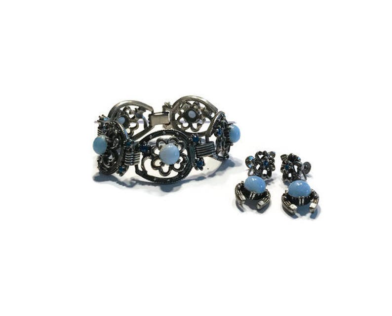 K&ouml;nnte beinhalten: Ein silberfarbenes Armband und ein passendes Ohrring-Set. Das Armband hat offene florale Designs mit hellblauen Cabochon-Steinen. Die Ohrringe haben ein &auml;hnliches Design mit kleineren blauen Steinen.