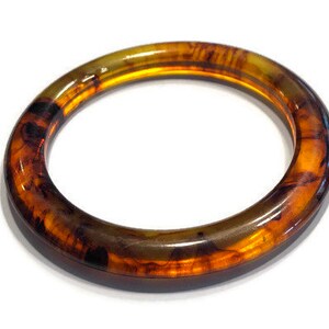 May include: A circular bangle bracelet featuring a translucent amber and dark brown tortoiseshell pattern. The accessory has a smooth, polished finish with a rounded profile, showcasing deep golden and dark brown swirls throughout the material.