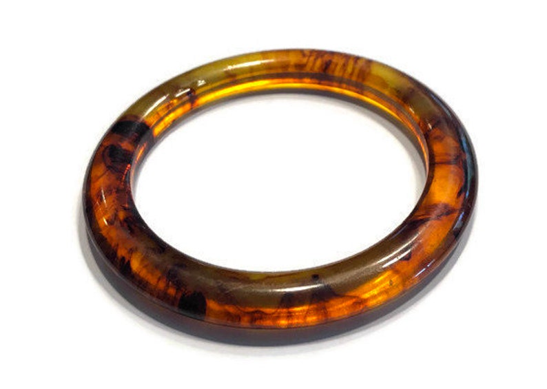 May include: A circular bangle bracelet featuring a translucent amber and dark brown tortoiseshell pattern. The accessory has a smooth, polished finish with a rounded profile, showcasing deep golden and dark brown swirls throughout the material.