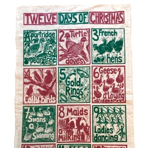 May include: A vintage-style tea towel with a red and green illustration of the Twelve Days of Christmas. The illustration features a different image for each day, such as a partridge in a pear tree, two turtle doves, and three French hens.
