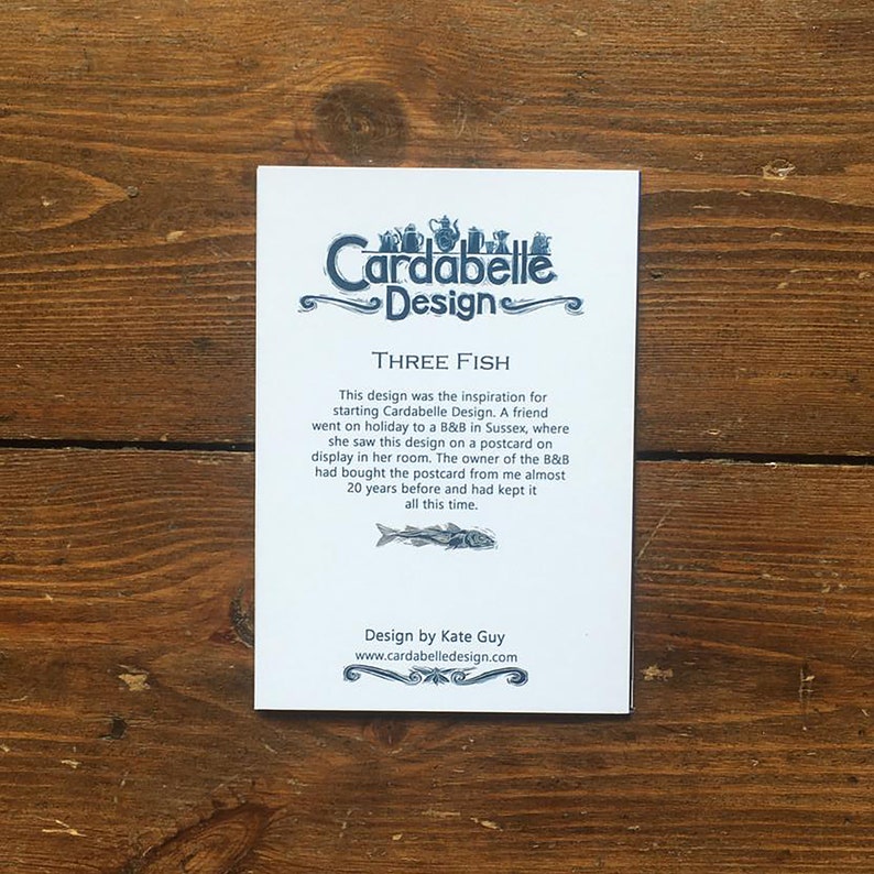 May include: A white card with blue text and a blue illustration of three fish. The text reads "Cardabelle Design" and "Three Fish". The text explains that the design was inspired by a postcard a friend saw on a trip to a B&B in Sussex. The text also says "Design by Kate Guy" and "www.cardabelledesign.com".