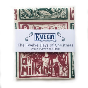 May include: A red and green tea towel with a vintage illustration of a person milking a cow. The towel is packaged and labeled "The Twelve Days of Christmas Organic Cotton Tea Towel" with the brand name "Kate Guy".
