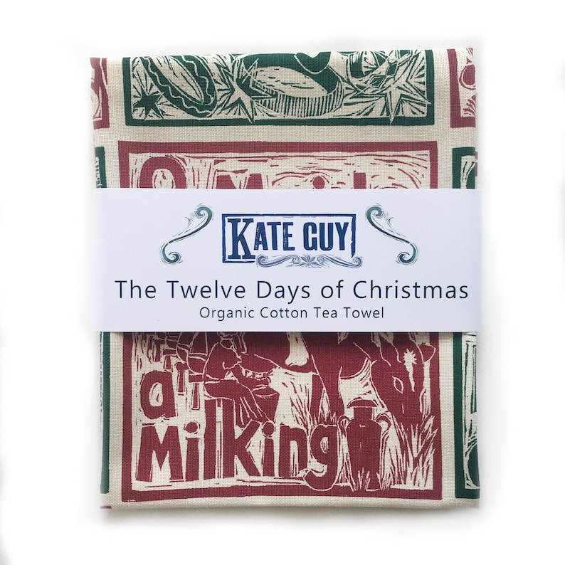 May include: A red and green tea towel with a vintage illustration of a person milking a cow. The towel is packaged and labeled "The Twelve Days of Christmas Organic Cotton Tea Towel" with the brand name "Kate Guy".