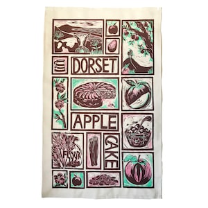 以下が含まれることがあります： リンゴ、シナモンスティック、小麦粉、そして「ドーセットアップルケーキ」の文字がブロックプリントされたデザインの茶色と緑のキッチンクロスです。