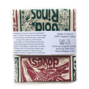 May include: A tea towel with a red and green linocut print of a bird and the words "Twelve Days of Christmas" in a stylized font. The text "Made in the UK 100% Organic Cotton" and "From an original linocut print by Kate Guy www.kateguy.co.uk" is printed on the bottom right corner of the towel.