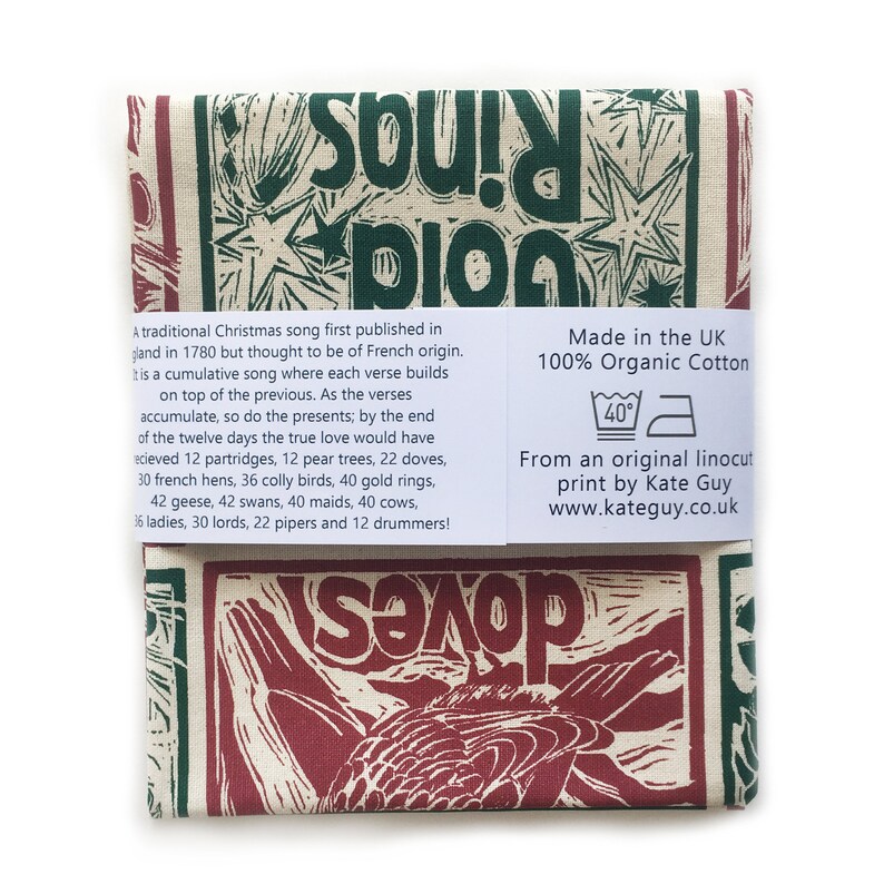 May include: A tea towel with a red and green linocut print of a bird and the words "Twelve Days of Christmas" in a stylized font. The text "Made in the UK 100% Organic Cotton" and "From an original linocut print by Kate Guy www.kateguy.co.uk" is printed on the bottom right corner of the towel.