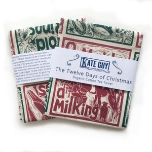 May include: A red and green tea towel with a block print design of the Twelve Days of Christmas song lyrics. The towel is made of organic cotton and has a white label with the text "The Twelve Days of Christmas Organic Cotton Tea Towel" and the brand name "KATE GUY".