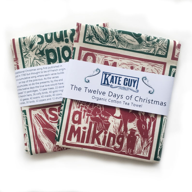 May include: A red and green tea towel with a block print design of the Twelve Days of Christmas song lyrics. The towel is made of organic cotton and has a white label with the text "The Twelve Days of Christmas Organic Cotton Tea Towel" and the brand name "KATE GUY".
