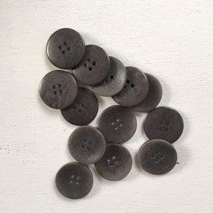 May include: A collection of 14 dark gray buttons with four holes each. The buttons are made of a textured material and have a slightly irregular shape.