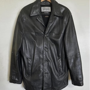 Puede incluir: Chaqueta de cuero negra con cierre de botones y cuello. La chaqueta tiene dos bolsillos delanteros y una etiqueta que dice "Pelle Studio". La chaqueta está colgada de una percha de madera.