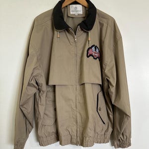 Peut inclure: Veste beige avec col noir et fermeture éclair. La veste présente un écusson avec un motif de drapeau américain. Elle possède deux poches avant et un cordon de serrage à la capuche. La veste est suspendue à un cintre en bois.