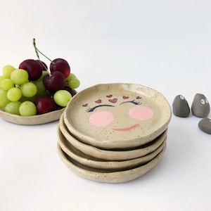 May include: A stack of speckled ceramic plates with a smiling face design. The face has pink cheeks, a red smiling mouth, and hearts above the eyes. A bowl of grapes and decorative stones are also in the image.
