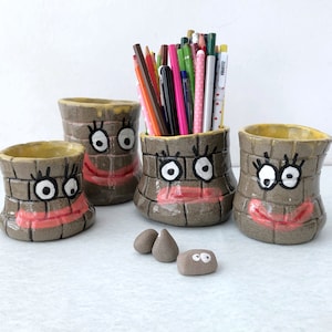 May include: Four whimsical ceramic pencil holders with cartoon faces, each with a brown brick-like pattern, yellow interior, and a pink mouth. One holder is filled with colorful pencils. Three small, gray, stone-like objects are in front of the holders.