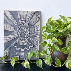 May include: A gray ceramic wall art piece featuring a clown with a hat and a radiant background. The clown has a detailed face and is wearing a ruffled collar. A potted plant with green and yellow leaves is next to the art.
