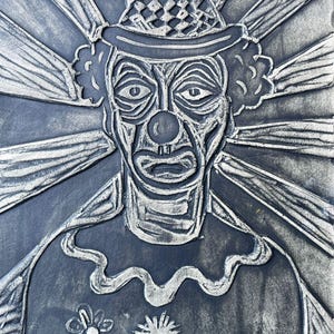 May include: A black and silver relief plaque featuring a central clown figure wearing a hat with a checkered pattern. The clown has a round nose, a ruffled collar, and a shirt decorated with two flower motifs. Radiating lines extend outward.