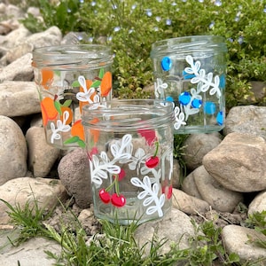 May include: Three glass jars with painted fruit designs. The jars are decorated with white leaves and orange slices, blueberries, and cherries.