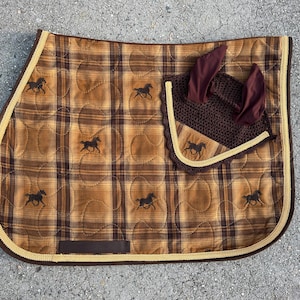 May include: A brown and tan plaid quilted saddle pad with a brown horse silhouette design. A brown and tan fly bonnet with a brown crochet mesh is attached to the saddle pad.