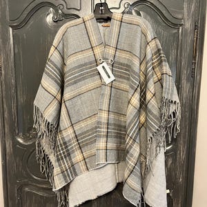 May include: A gray and beige plaid poncho with fringe trim. The poncho has a metal ring detail and a tag that reads "Made in Italy".