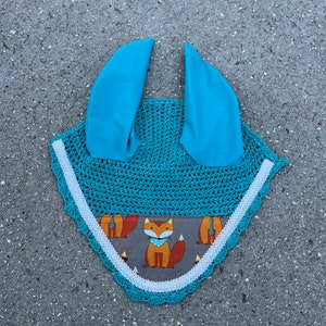 May include: A turquoise crochet horse fly bonnet with white lace trim and a fox print fabric panel. The bonnet has two ear pieces.
