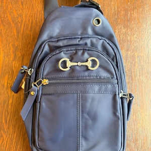 May include: Navy blue sling bag with a gold-colored snaffle bit detail on the front. The bag has a zippered main compartment and a smaller front pocket. It features a single adjustable shoulder strap and a small handle at the top.
