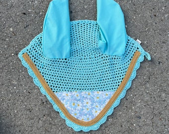 May include: A light blue crochet horse bonnet with matching ear covers. The bonnet features a gold trim and a floral daisy pattern. This equestrian accessory is designed to protect a horse's ears from insects and noise.