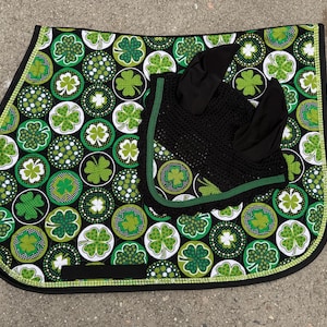 May include: A black and green equestrian saddle pad and matching ear bonnet set. The saddle pad features a pattern of green shamrocks on a black background, with a green and white border. The ear bonnet is black with green trim.