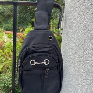 May include: Black crossbody bag with a silver-toned metal accent featuring a design of two interlocking circles with rhinestones. The bag has a zippered pocket and a strap.