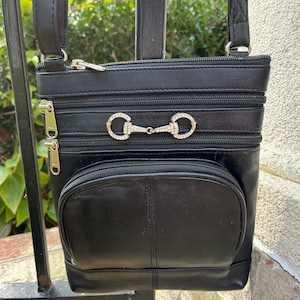 May include: Black leather crossbody bag with a silver and rhinestone horse bit detail. The bag has multiple zippered compartments.