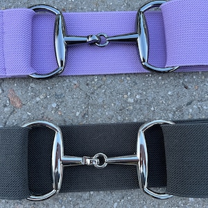 May include: Two elastic belts with silver-toned metal buckles. The top belt is lavender and the bottom belt is black.