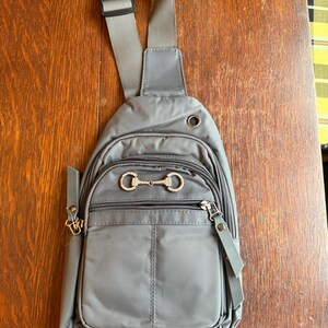 May include: Gray sling bag with a single adjustable shoulder strap. The bag features multiple zippered compartments and a decorative silver-colored horse bit detail on the front. The bag is made of a smooth, durable material.
