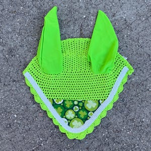 May include: A neon green crochet horse bonnet with two upright ear covers. The bonnet has a white trim and a green and white clover pattern. The item is designed to protect a horse's ears from insects and noise.