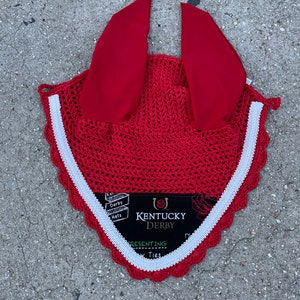 May include: A red crochet horse bonnet with matching red ear covers and a white trim. The bonnet has a scalloped edge and a tag that reads "Kentucky Derby".