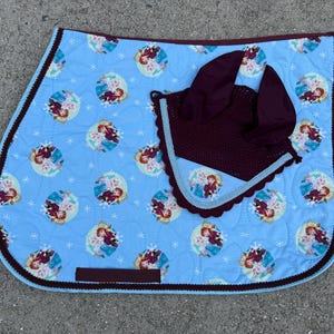 May include: A light blue quilted saddle pad with a repeating pattern of cartoon characters in a circular design. A matching burgundy crochet bonnet and trim complete the set. The pad has a burgundy border.