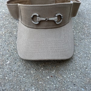 May include: A tan visor with a silver bit design embellished with clear rhinestones. The visor has a curved brim and a fabric band. The visor is made of a woven material.
