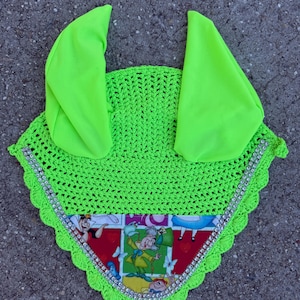 May include: A neon green crochet horse bonnet with ear covers. The bonnet features a colorful Alice in Wonderland print along the bottom edge, embellished with silver-colored rhinestones. The ear covers are made of a solid neon green fabric.