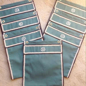 Può includere: Un set di 12 borse in tela color teal con cuciture bianche e bordi neri. Ogni borsa ha un monogramma ricamato bianco sul davanti.