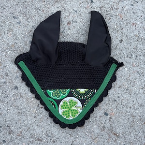 May include: Black crochet horse bonnet with black ear covers and a green and white clover pattern. The bonnet has a green scalloped trim and is designed to protect a horse's ears from insects and noise.