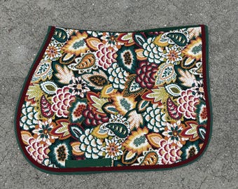 May include: A colorful saddle pad with a floral pattern in shades of yellow, orange, red, and green. The pad has a dark green border with a maroon trim. The design features stylized flowers and paisley shapes.